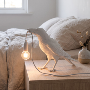 White raven lamp lit on a bedside table in a bright bedroom